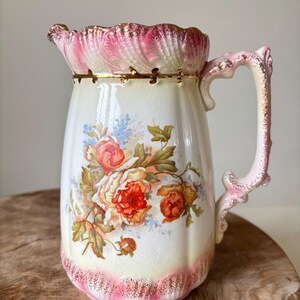 Peut inclure: Pichet en céramique blanche vintage avec un bord et une anse festonnés roses et dorés. Le pichet est décoré d'un motif floral avec des roses rouges et oranges et des feuilles vertes. La base du pichet est également ornée de rose et d'or.