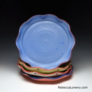 May include: A stack of four ceramic plates with scalloped edges. The top plate is light blue. The lower plates are yellow, white, and red, each featuring small blue and red circular accents along the rim. The text RebeccaLowery.com is visible.