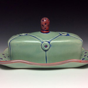 May include: A green ceramic butter dish with a red knob on the lid. The dish has a scalloped edge and is decorated with blue and red circles.