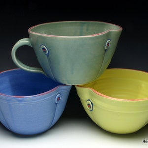 Może przedstawiać: Trzy ceramiczne miski do mieszania z uchwytami. Miski są w odcieniach niebieskiego, zielonego i żółtego. Każda miska ma czerwoną krawędź i ozdobny wzór w postaci koła z czerwonym punktem w środku.