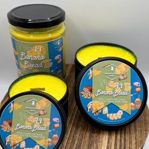 May include: A selection of candles with yellow wax and black lids. The labels read "Chapter 14 Banana Bread" with ingredients listed. One candle is in a glass jar, and the others are in black tins. The candles are presented on a wooden board.