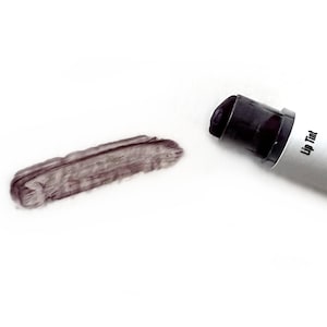 Könnte beinhalten: Eine Tube Raven Wine Lippenfarbe mit einem dunkelvioletten Applikator und einer Produktprobe. Die Tube ist silberfarben mit schwarzem Text, der "SIMPLE Traditions" und "fresh handcrafted luxury" lautet. Das Produkt hat 2 ml.