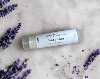May include: A clear lip balm tube with a white label that reads "Lavender" and "fresh handcrafted beauty" is surrounded by sprigs of purple lavender flowers. The background is a light gray, textured surface.