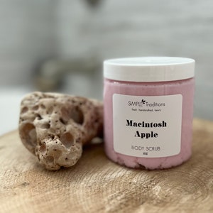 May include: A jar of pink body scrub labeled "Macintosh Apple" sits on a wooden surface next to a porous, beige stone. The jar has a white lid and the text "SIMPLE Traditions" is above the product name.