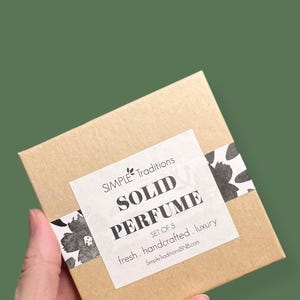 May include: A brown cardboard box with a white label that says "SIMPLE Traditions SOLID PERFUME SET OF 5 fresh. handcrafted. luxury SimpleTraditionsBNB.com". The box is decorated with black and white floral designs.