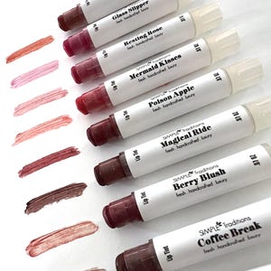 Puede incluir: Diez bálsamos labiales en tubos con etiquetas blancas y texto negro. Las etiquetas enumeran los nombres de los bálsamos labiales, que incluyen "Glass Slipper", "Resting Rose", "Mermaid Kisses", "Poison Apple", "Magical Ride", "Berry Blush", "Coffee Break" y "Berry Wine". Los bálsamos labiales están dispuestos en una fila del más claro al más oscuro, con el bálsamo más claro en la parte superior de la imagen y el bálsamo más oscuro en la parte inferior.