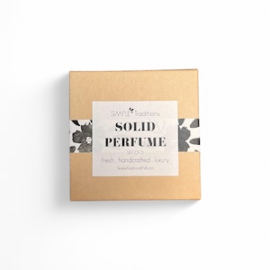 May include: A brown cardboard box with a white label that reads "SIMPLE Traditions SOLID PERFUME SET OF 5 fresh. handcrafted. luxury SimpletraditionsBN8.com". The box has a black and white floral design on the top and bottom.