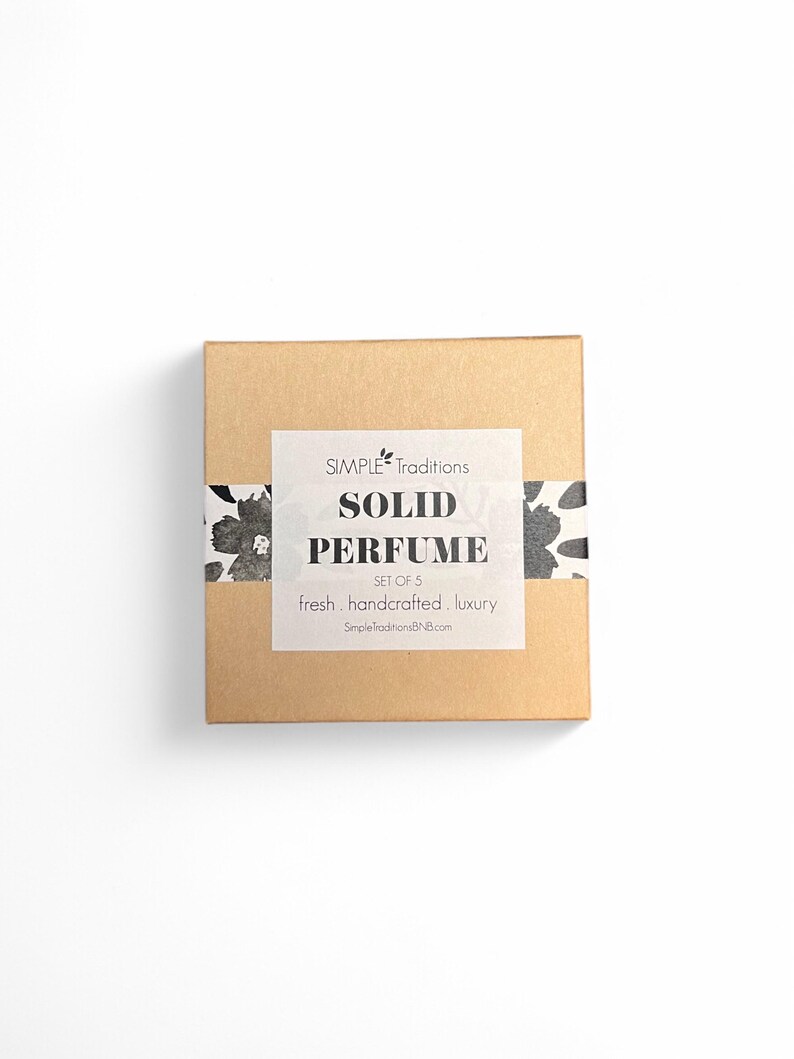 May include: A brown cardboard box with a white label that reads "SIMPLE Traditions SOLID PERFUME SET OF 5 fresh. handcrafted. luxury SimpletraditionsBN8.com". The box has a black and white floral design on the top and bottom.
