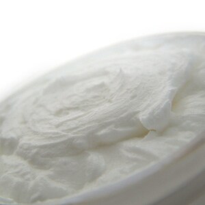 May include: A thick, white, opaque cream with a smooth, whipped texture contained within a round jar. The surface of the cream displays soft peaks and swirls, indicating a dense, rich consistency that fills the entire circular opening of the container.