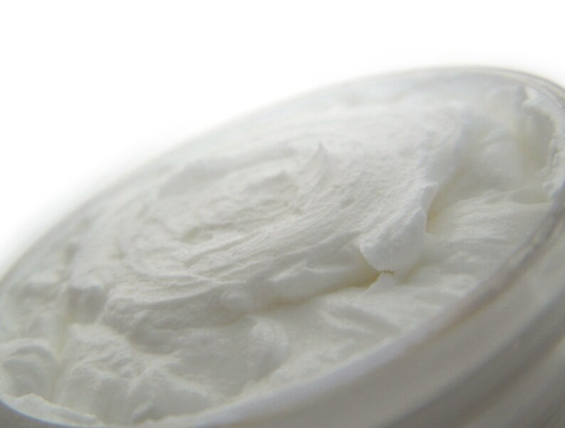 May include: A thick, white, opaque cream with a smooth, whipped texture contained within a round jar. The surface of the cream displays soft peaks and swirls, indicating a dense, rich consistency that fills the entire circular opening of the container.