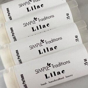 Pu&ograve; includere: Quattro tubetti bianchi di profumo solido con etichette nere. Le etichette riportano "SIMPLE Traditions Lilac" e ".15 oz". L'etichetta inferiore riporta anche "fresh. handcrafted. luxury".
