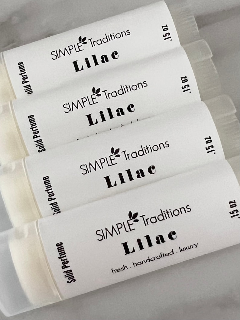 Pu&ograve; includere: Quattro tubetti bianchi di profumo solido con etichette nere. Le etichette riportano "SIMPLE Traditions Lilac" e ".15 oz". L'etichetta inferiore riporta anche "fresh. handcrafted. luxury".