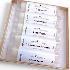 May include: Set of five small clear tubes containing solid perfume. Each tube has a white label with the brand name "SIMPLE Traditions" and a different scent name: "Romance", "Enchanting", "Captivate", "Bohemian Nectar", and "First Kiss". Each label also includes the text "fresh handcrafted luxury" and the size ".15 oz".