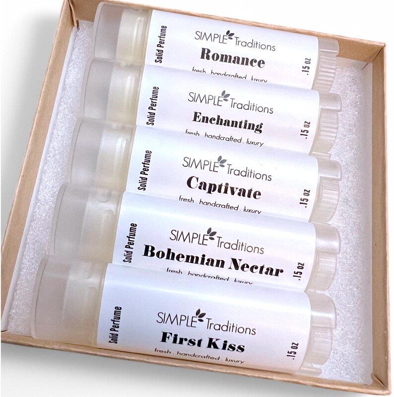 May include: Set of five small clear tubes containing solid perfume. Each tube has a white label with the brand name "SIMPLE Traditions" and a different scent name: "Romance", "Enchanting", "Captivate", "Bohemian Nectar", and "First Kiss". Each label also includes the text "fresh handcrafted luxury" and the size ".15 oz".