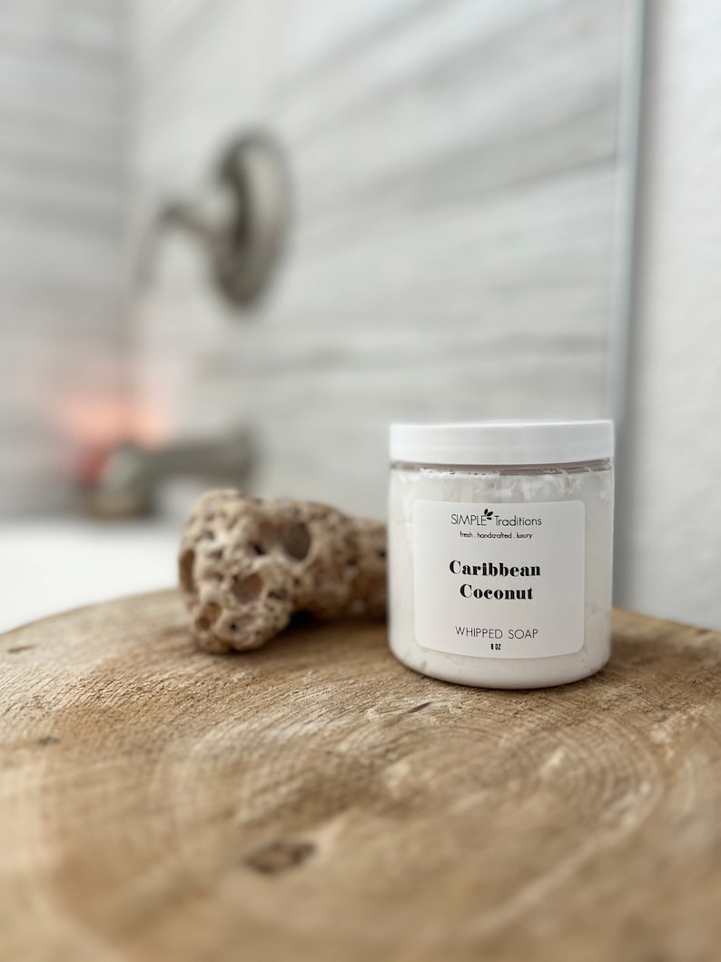 May include: A clear plastic jar filled with white whipped soap, topped with a white lid. The label reads Simple Traditions, Caribbean Coconut, and WHIPPED SOAP 8 oz. The jar is positioned next to a porous, light brown textured stone.