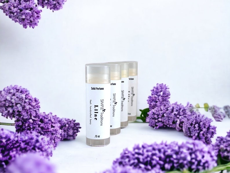 Pu&ograve; includere: Tre piccoli tubetti di profumo solido bianchi con il testo "SIMPLE Traditions Lilac" sul davanti. I tubetti sono disposti in fila su una superficie bianca con fiori di lill&agrave; viola sullo sfondo.