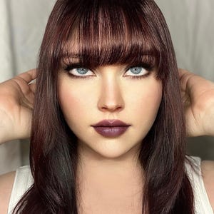 K&ouml;nnte beinhalten: Portr&auml;t einer Person mit langem, dunkelbraunem Haar und Pony. Die Person hat hellblaue Augen, dunkles Augen-Make-up und dunkelvioletten Lippenstift. Die Person tr&auml;gt ein wei&szlig;es Oberteil.