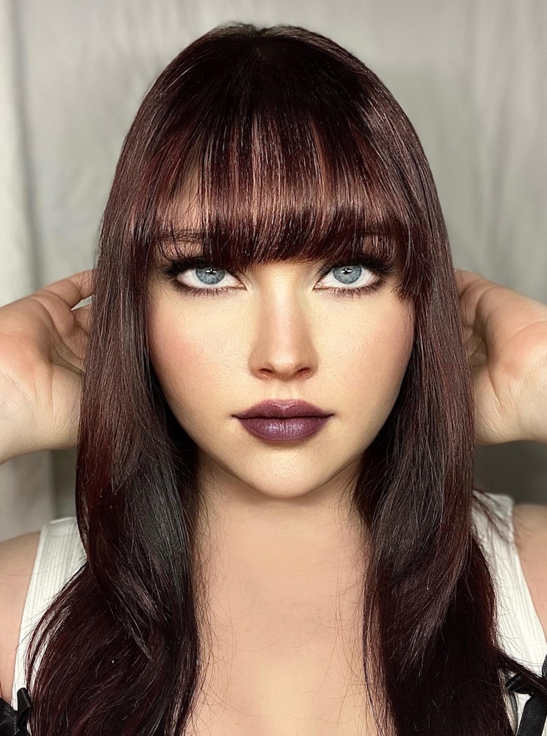 K&ouml;nnte beinhalten: Portr&auml;t einer Person mit langem, dunkelbraunem Haar und Pony. Die Person hat hellblaue Augen, dunkles Augen-Make-up und dunkelvioletten Lippenstift. Die Person tr&auml;gt ein wei&szlig;es Oberteil.