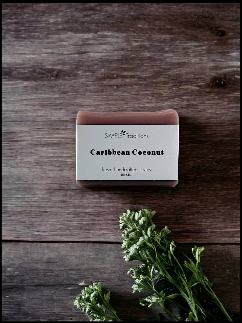 May include: A bar of brown soap with a white label that reads "SIMPLE Traditions Caribbean Coconut fresh. handcrafted. luxury. SOAP 4.5 OZ". The soap is sitting on a wooden surface with green plants in the foreground.
