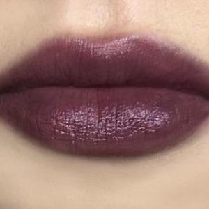K&ouml;nnte beinhalten: Nahaufnahme von Lippen, die mit einem tiefen Pflaumen-Lippenstift geschminkt sind. Die Farbe ist reichhaltig und gl&auml;nzend, mit leichtem Schimmer. Die Lippen sind voll und gut definiert, mit einer glatten Textur. Die Haut um die Lippen ist hell.