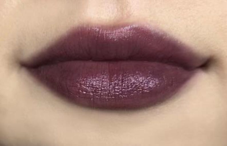 K&ouml;nnte beinhalten: Nahaufnahme von Lippen, die mit einem tiefen Pflaumen-Lippenstift geschminkt sind. Die Farbe ist reichhaltig und gl&auml;nzend, mit leichtem Schimmer. Die Lippen sind voll und gut definiert, mit einer glatten Textur. Die Haut um die Lippen ist hell.