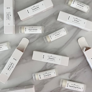 May include: A variety of lip balm tubes with different scents, including Gardenia, Jasmine, Lilac, Nag Champa, Captivate, First Kiss, and Sparkling Berry. The lip balms are packaged in white boxes with black text.