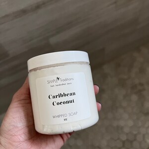 May include: A white plastic jar of whipped soap with a white screw-top lid. The label reads Simple Traditions, fresh handcrafted luxury, Caribbean Coconut, WHIPPED SOAP, and 8 OZ. The container is filled with a white, creamy substance.