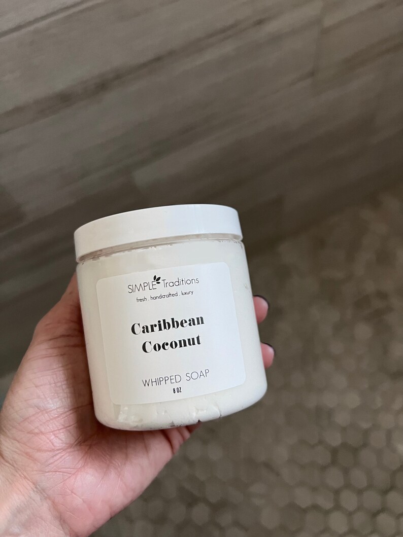 May include: A white plastic jar of whipped soap with a white screw-top lid. The label reads Simple Traditions, fresh handcrafted luxury, Caribbean Coconut, WHIPPED SOAP, and 8 OZ. The container is filled with a white, creamy substance.