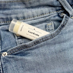 Puede incluir: Un stick de perfume sólido blanco y transparente etiquetado como "SIMPLE Traditions Gardenia" está metido en un bolsillo de unos vaqueros de mezclilla. La etiqueta también dice "fresh, handcrafted luxury". El perfume tiene un tono amarillo claro.