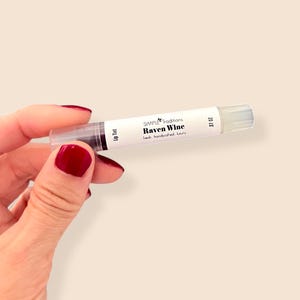 K&ouml;nnte beinhalten: Eine transparente Lip-Tint-Tube mit der Aufschrift "Raven Wine" von Simple Traditions. Die Tube enth&auml;lt ein dunkles, weinfarbenes Produkt und hat eine transparente Applikatorspitze. Der Text "fresh handcrafted luxury" ist ebenfalls aufgedruckt.
