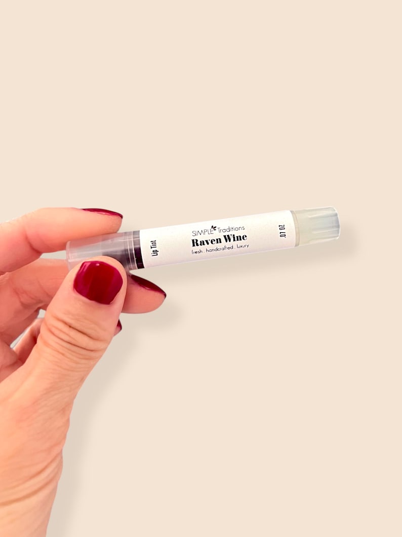 K&ouml;nnte beinhalten: Eine transparente Lip-Tint-Tube mit der Aufschrift "Raven Wine" von Simple Traditions. Die Tube enth&auml;lt ein dunkles, weinfarbenes Produkt und hat eine transparente Applikatorspitze. Der Text "fresh handcrafted luxury" ist ebenfalls aufgedruckt.