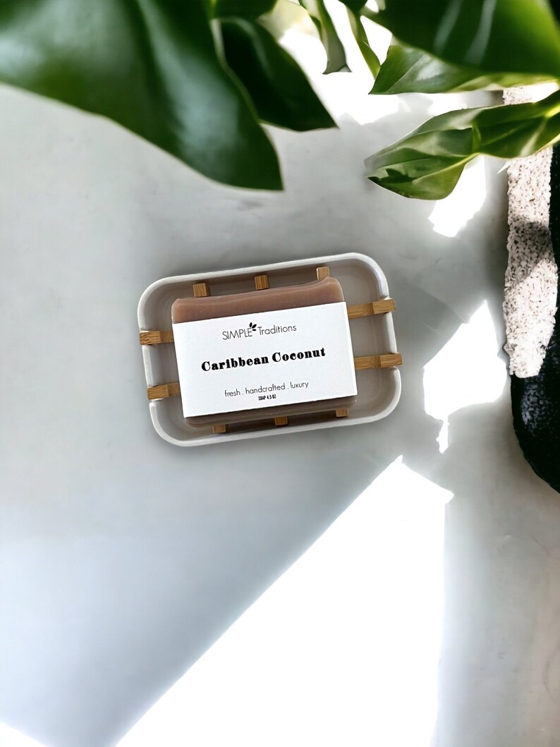 May include: A bar of brown soap in a white dish with a bamboo soap dish. The soap is labeled "SIMPLE Traditions Caribbean Coconut fresh, handcrafted, luxury".