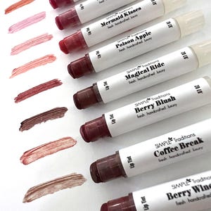 K&ouml;nnte beinhalten: Eine Auswahl an Lippenfarben in verschiedenen Farbt&ouml;nen, von Rosa bis Braun, mit passenden Farbmustern. Jede Tube ist mit Namen wie "Glass Slipper" und "Coffee Break" beschriftet. Der Markenname "SIMPLE Traditions" ist sichtbar.