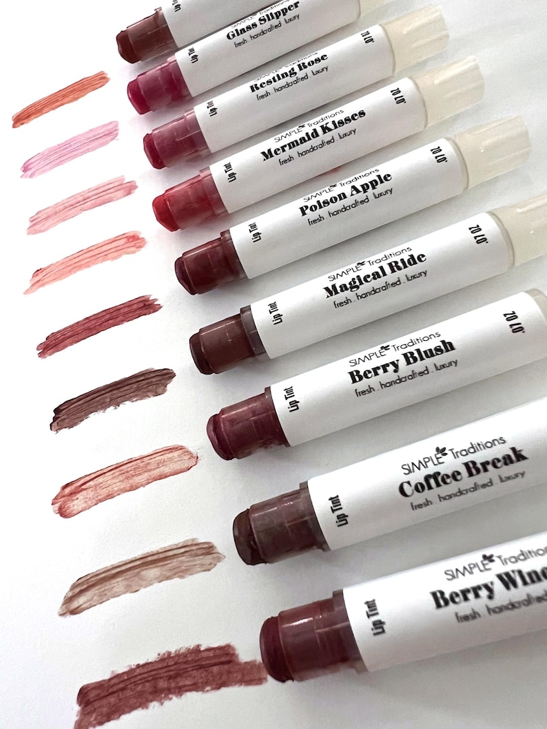 K&ouml;nnte beinhalten: Eine Auswahl an Lippenfarben in verschiedenen Farbt&ouml;nen, von Rosa bis Braun, mit passenden Farbmustern. Jede Tube ist mit Namen wie "Glass Slipper" und "Coffee Break" beschriftet. Der Markenname "SIMPLE Traditions" ist sichtbar.