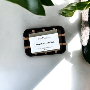 May include: A bar of green clay soap in a black and bamboo soap dish. The soap label reads "SIMPLE traditions French Green Clay fresh handcrafted luxury".