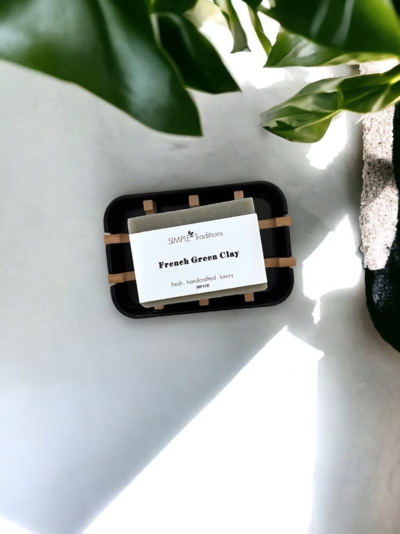 May include: A bar of green clay soap in a black and bamboo soap dish. The soap label reads "SIMPLE traditions French Green Clay fresh handcrafted luxury".