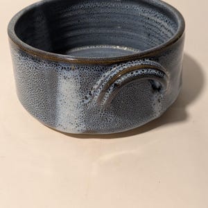 May include: A small, blue ceramic bowl with a speckled glaze and a brown rim. The bowl has two handles on the sides.