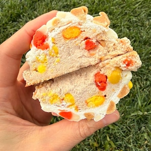 Könnte beinhalten: Ein angeschnittenes Dessert, gehalten in einer Hand, zeigt Schichten aus hellbraunen, weißen und bunten Süßigkeiten. Das Dessert hat eine weiche Textur mit sichtbaren orangefarbenen, gelben und roten Süßigkeiten und ist mit kleinen, hellen Stücken belegt.