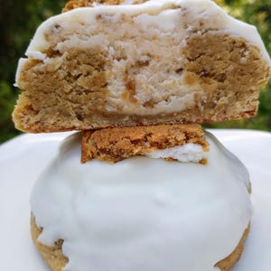 May include: A close-up of a cookie with a white frosting and a piece of cookie on top. The cookie is cut in half, revealing a creamy filling. The top of the cookie has a crumbly topping. The cookie is on a white plate.