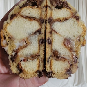 May include: A cross-section of a baked pastry, revealing layers of bread, chocolate chips, and a sweet glaze. The pastry is held in a hand, showcasing the texture and ingredients. The dessert appears to be a type of bread pudding or similar treat.