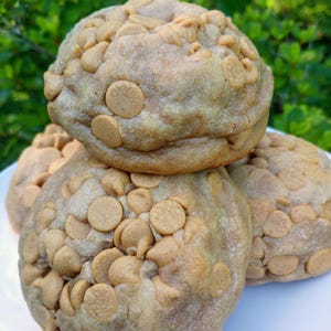 Peut inclure: Quatre gros cookies ronds, de couleur brun clair, empilés sur une assiette blanche. Les cookies sont parsemés de pépites de beurre de cacahuète. L'arrière-plan est un feuillage vert flou.