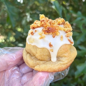 May include: A round, golden-brown baked cookie topped with a thick layer of smooth white icing and a generous sprinkling of toasted, crunchy golden-brown crumbles. The cookie has a soft, rounded shape and a visible drip of icing down the side.