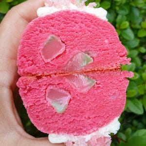 May include: A pink dessert cut in half, revealing jelly pieces. The dessert has a textured, cake-like appearance, with white frosting and pink and green sugar crystals on top. The dessert is held in a hand against a backdrop of green leaves.