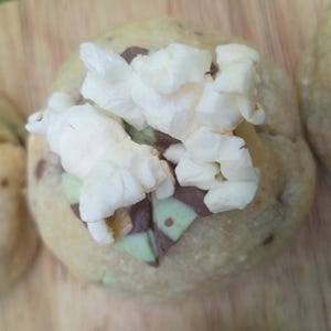 May include: A round, golden-brown baked cookie topped with white popcorn kernels and small, triangular pieces of mint-green and dark brown chocolate. The cookie has a textured surface with visible dark brown inclusions embedded within the dough.