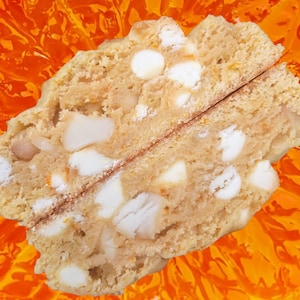 May include: A close-up of a biscotti cut in half, revealing a light brown interior studded with white chocolate chips. The biscotti sits against an orange, abstract background.
