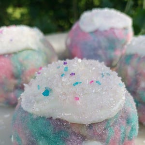 May include: Close-up of four colorful, frosted treats. Each treat has a base of pastel colors, including pink, blue, and purple, covered in white frosting and sprinkled with colorful sugar crystals. The treats are arranged on a white surface.