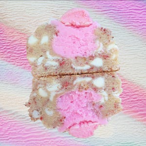 May include: A biscuity treat, possibly a cookie or scone, cut in half to reveal a pink filling and white chocolate chips. The background is a blend of pink and white, creating a soft, colourful backdrop for the baked good.