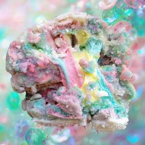 Op de afbeelding: Een close-up van een kleurrijk, gelaagd gebak. Het dessert heeft pastelroze, blauwe, gele en groene lagen, bestrooid met witte suikerkristallen. Het algehele effect is grillig en levendig, met een zachte, getextureerde uitstraling.