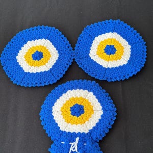 Op de afbeelding: Drie blauwe, witte en gele gehaakte onderzetters in de vorm van een oog. De onderzetters zijn gerangschikt op een zwart oppervlak. Het oogontwerp heeft een blauwe buitenring, een witte middenring en een gele kern.