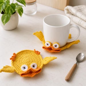 May include: Two round, yellow crocheted coasters shaped like ducks, each featuring orange wings, an orange beak, and white eyes with black cross-stitched pupils. One coaster sits flat, while the other supports a plain white ceramic mug.